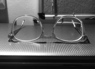 A pair of transparent eyeglasses rests on a textured surface, with a background featuring a blurred wall and part of a corkboard. The glasses have a square-shaped frame and cast a subtle shadow on the surface.
