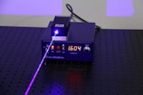 A black electronic device with a digital display showing the number 1.604, surrounded by control buttons. A purple laser beam is emitted from the device, projecting onto a perforated black surface.