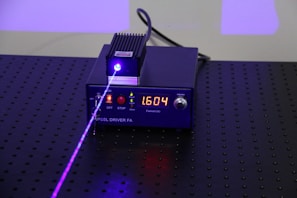 A black electronic device with a digital display showing the number 1.604, surrounded by control buttons. A purple laser beam is emitted from the device, projecting onto a perforated black surface.
