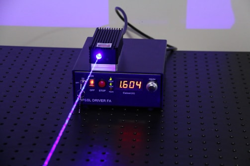 A black electronic device with a digital display showing the number 1.604, surrounded by control buttons. A purple laser beam is emitted from the device, projecting onto a perforated black surface.