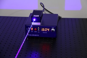 A black electronic device with a digital display showing the number 1.604, surrounded by control buttons. A purple laser beam is emitted from the device, projecting onto a perforated black surface.
