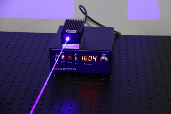 A black electronic device with a digital display showing the number 1.604, surrounded by control buttons. A purple laser beam is emitted from the device, projecting onto a perforated black surface.
