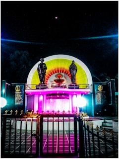 A monument features two statues on a platform, with vibrant lighting in purple, yellow, and red hues. The statues are positioned in front of a circular backdrop with multicolored sections. In the foreground, there is a black metal gate with golden spikes. The scene is set against a dark night sky.
