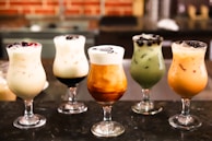 Five glasses of varying iced drinks are arranged in a row on a dark surface. Each glass contains a different type of creamy, colorful beverage, some with toppings like berries and boba pearls. The drinks are set against a blurred background, possibly a café setting.