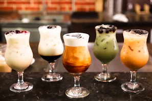 Five glasses of varying iced drinks are arranged in a row on a dark surface. Each glass contains a different type of creamy, colorful beverage, some with toppings like berries and boba pearls. The drinks are set against a blurred background, possibly a café setting.