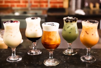 Five glasses of varying iced drinks are arranged in a row on a dark surface. Each glass contains a different type of creamy, colorful beverage, some with toppings like berries and boba pearls. The drinks are set against a blurred background, possibly a café setting.
