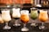 Five glasses of varying iced drinks are arranged in a row on a dark surface. Each glass contains a different type of creamy, colorful beverage, some with toppings like berries and boba pearls. The drinks are set against a blurred background, possibly a café setting.