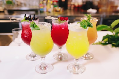 A selection of vibrantly colored tropical cocktails served in elegant glassware, each garnished with fresh mint leaves and decorative skewers. The drinks range in color from yellow to red, showcasing a refreshing appearance. They are placed on a white surface with a blurred background suggesting a bar setting.