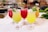A selection of vibrantly colored tropical cocktails served in elegant glassware, each garnished with fresh mint leaves and decorative skewers. The drinks range in color from yellow to red, showcasing a refreshing appearance. They are placed on a white surface with a blurred background suggesting a bar setting.