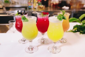 A selection of vibrantly colored tropical cocktails served in elegant glassware, each garnished with fresh mint leaves and decorative skewers. The drinks range in color from yellow to red, showcasing a refreshing appearance. They are placed on a white surface with a blurred background suggesting a bar setting.