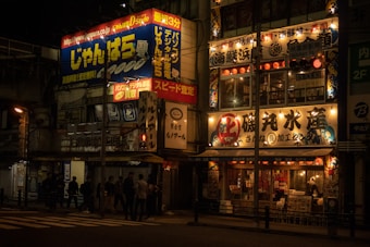 A nighttime urban scene with brightly lit Japanese storefronts and signage, featuring colorful advertising and illuminated text in Japanese characters. People are walking along the sidewalk under the glow of streetlights, and the atmosphere is bustling and lively.