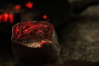A close-up view of a reflective metallic container, potentially made of foil, with small perforations and glowing red embers inside. The background is dark and blurry, with a few red glowing points of light.