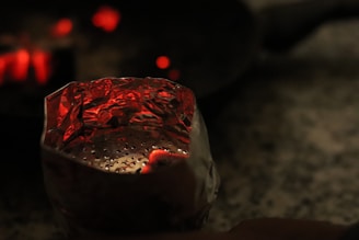 A close-up view of a reflective metallic container, potentially made of foil, with small perforations and glowing red embers inside. The background is dark and blurry, with a few red glowing points of light.