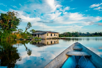 A serene river scene with a wooden house situated on the riverbank, surrounded by lush greenery and trees. In the foreground, the bow of a boat points towards the house. The sky is blue with scattered clouds, and the water reflects the landscape and sky, creating a mirror-like surface.