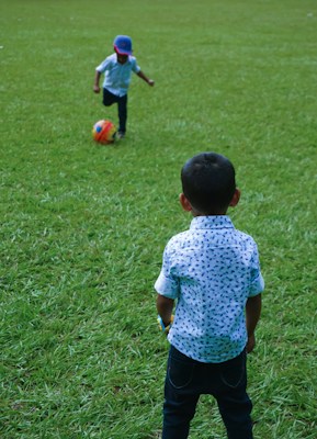 Two young children are playing with a colorful soccer ball on a grassy field. One child is in the foreground with his back to the camera, wearing a patterned shirt, while the other child is running towards the ball, wearing a cap.