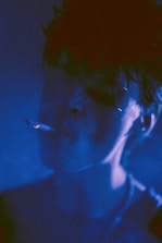 A person with a cigarette in their mouth is partially illuminated by a cool blue light, creating a moody and atmospheric effect. The lighting obscures much of the facial detail, highlighting the contours of the face and the smoke emanating from the cigarette.