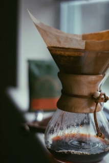 A high-quality coffee maker with a wooden neck and leather strap is positioned near a window. The glass carafe is partially filled with brewed coffee, and a paper filter holds the coffee grounds on top. The background features a blurry view, possibly of another object or packaging.