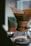 A high-quality coffee maker with a wooden neck and leather strap is positioned near a window. The glass carafe is partially filled with brewed coffee, and a paper filter holds the coffee grounds on top. The background features a blurry view, possibly of another object or packaging.