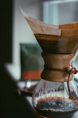 A high-quality coffee maker with a wooden neck and leather strap is positioned near a window. The glass carafe is partially filled with brewed coffee, and a paper filter holds the coffee grounds on top. The background features a blurry view, possibly of another object or packaging.