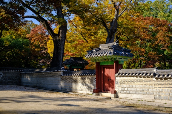 A traditional Korean stone wall and wooden gate with a tiled roof stands against a backdrop of lush autumn trees with golden and red leaves.