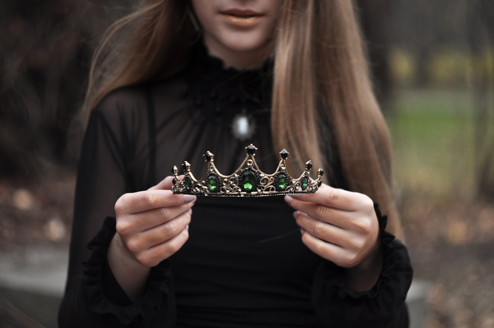 girl wearing black long-sleeved blouse standing while holding green and silver tiara