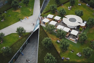 An aerial view of a public park featuring a geometric design with intersecting pathways, grassy areas, and scattered trees. A circular structure with several canopies serves as a seating and dining area. People are walking, sitting, and enjoying the outdoor space.