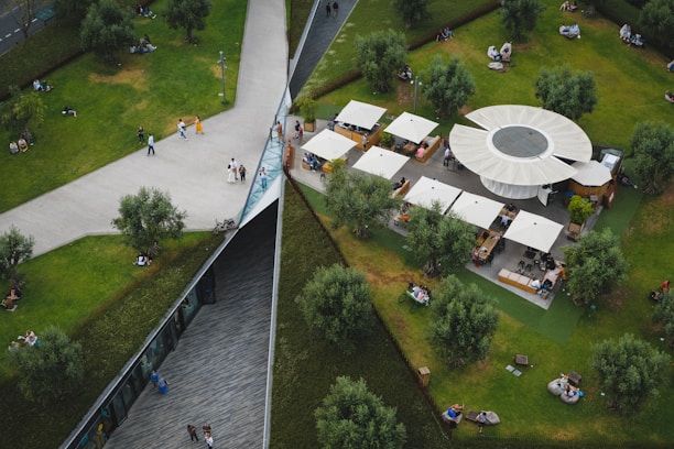 An aerial view of a public park featuring a geometric design with intersecting pathways, grassy areas, and scattered trees. A circular structure with several canopies serves as a seating and dining area. People are walking, sitting, and enjoying the outdoor space.