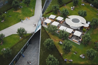 An aerial view of a public park featuring a geometric design with intersecting pathways, grassy areas, and scattered trees. A circular structure with several canopies serves as a seating and dining area. People are walking, sitting, and enjoying the outdoor space.