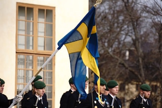 A group of uniformed individuals, possibly soldiers, stand in formation outdoors. A prominent Swedish flag is being held by one of the individuals. They are wearing green berets and appear to be participating in a formal or ceremonial event. A large windowed building and leafless trees are visible in the background.
