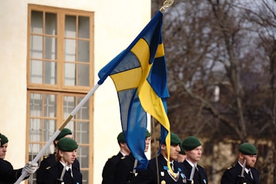 A group of uniformed individuals, possibly soldiers, stand in formation outdoors. A prominent Swedish flag is being held by one of the individuals. They are wearing green berets and appear to be participating in a formal or ceremonial event. A large windowed building and leafless trees are visible in the background.