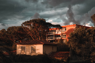 A residential area featuring a small, pale yellow house with closed shutters and a red-tiled roof is set against a backdrop of tall pine trees and a larger apartment building. The warm glow of late-afternoon sunlight contrasts with the dark, dramatic clouds looming overhead, casting an atmospheric light on the scene.
