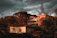 A residential area featuring a small, pale yellow house with closed shutters and a red-tiled roof is set against a backdrop of tall pine trees and a larger apartment building. The warm glow of late-afternoon sunlight contrasts with the dark, dramatic clouds looming overhead, casting an atmospheric light on the scene.