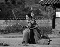 A person in traditional warrior attire is kneeling on one knee while wielding a sword with an outstretched arm. The background features a stone wall and traditional tiled roof structure. The presence of foliage and dirt ground suggests an outdoor setting.