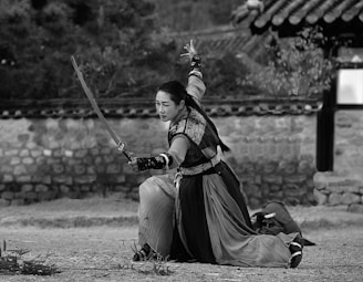 A person in traditional warrior attire is kneeling on one knee while wielding a sword with an outstretched arm. The background features a stone wall and traditional tiled roof structure. The presence of foliage and dirt ground suggests an outdoor setting.