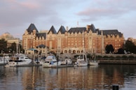 A grand, historic hotel with a distinct architectural style featuring steeply pitched roofs, multiple dormer windows, and ornate detailing. In front, a serene harbor bordered by trees and buildings hosts several yachts and boats gently floating on the water, reflecting the building's light.