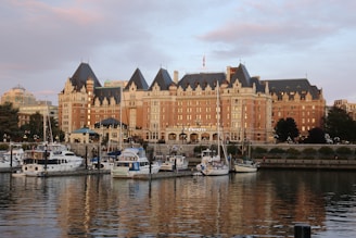 A grand, historic hotel with a distinct architectural style featuring steeply pitched roofs, multiple dormer windows, and ornate detailing. In front, a serene harbor bordered by trees and buildings hosts several yachts and boats gently floating on the water, reflecting the building's light.