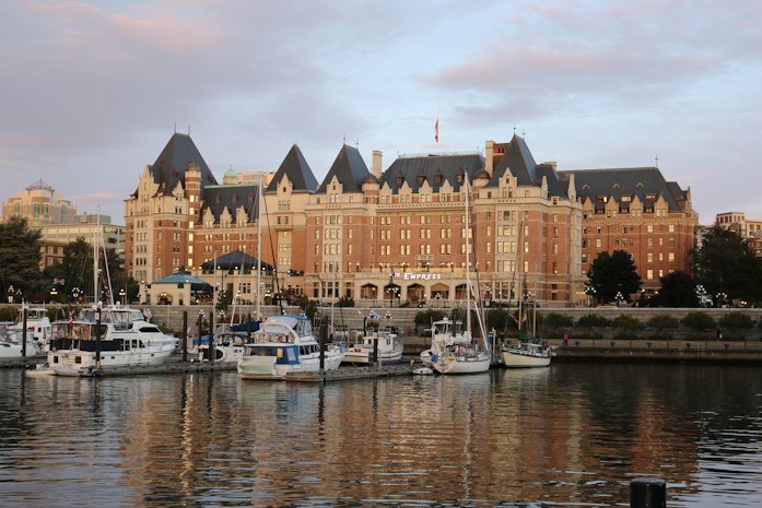 A grand, historic hotel with a distinct architectural style featuring steeply pitched roofs, multiple dormer windows, and ornate detailing. In front, a serene harbor bordered by trees and buildings hosts several yachts and boats gently floating on the water, reflecting the building's light.