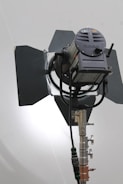 A large professional film or photography studio light with adjustable barn doors and a sturdy metal stand. The light source appears to be positioned against a plain background with soft illumination.