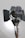 A large professional film or photography studio light with adjustable barn doors and a sturdy metal stand. The light source appears to be positioned against a plain background with soft illumination.
