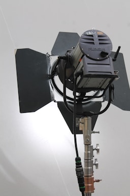 A large professional film or photography studio light with adjustable barn doors and a sturdy metal stand. The light source appears to be positioned against a plain background with soft illumination.