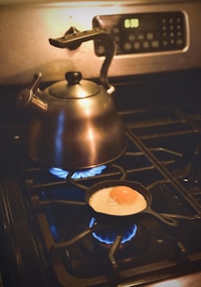 A black tea kettle sits on a stove burner with a blue flame underneath, while a small frying pan beside it cooks a sunny-side-up egg over another lit burner. An oven clock displays the time as 6:30.