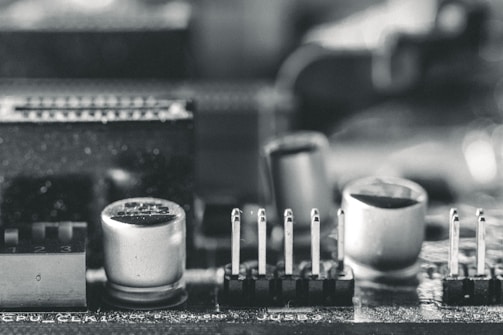 A macro close-up of an electronic circuit board featuring components such as capacitors, pins, and connectors. The image is in black and white, highlighting the intricate details and textures of the hardware elements.