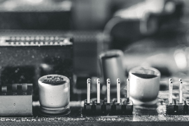 A macro close-up of an electronic circuit board featuring components such as capacitors, pins, and connectors. The image is in black and white, highlighting the intricate details and textures of the hardware elements.