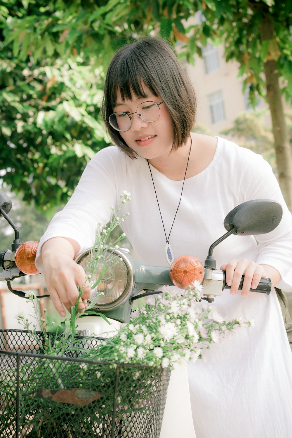 smiling woman wearing white crew-neck long-sleeved dress and eyeglasses standing near teal motor scooter