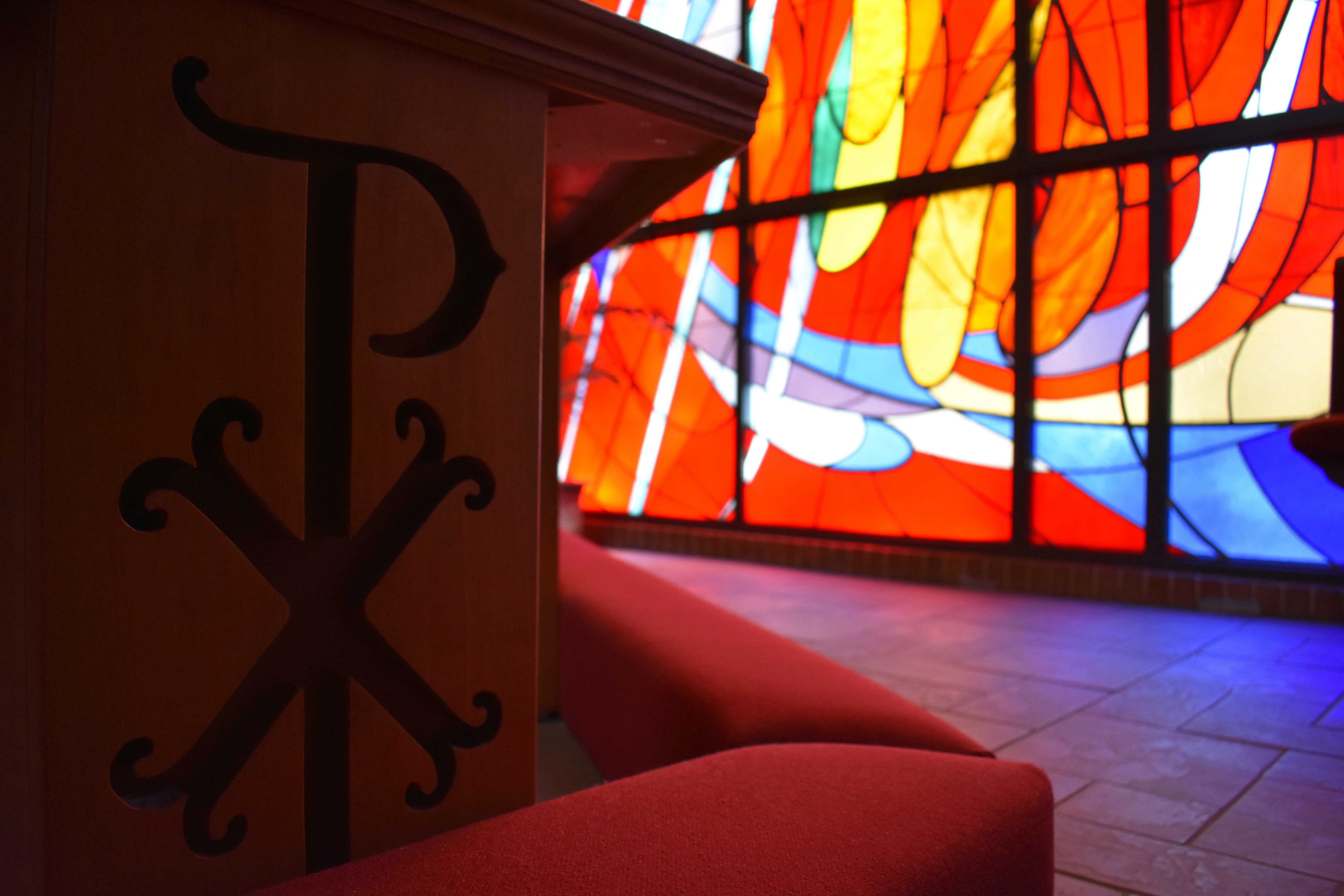 A close-up on a Chi-Rho in our communion railing, with one of our stained-glass windows behind.