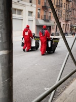 Two people dressed in red protective uniforms are pushing large black garbage bins on a city street. The background features urban architecture with brick buildings and some metal scaffolding in the foreground. The street appears relatively empty and the sky is not visible.