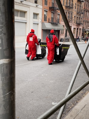 Two people dressed in red protective uniforms are pushing large black garbage bins on a city street. The background features urban architecture with brick buildings and some metal scaffolding in the foreground. The street appears relatively empty and the sky is not visible.