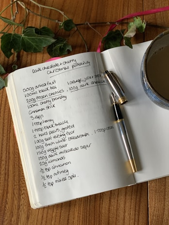 An open notebook displays a handwritten recipe for dark chocolate and cherry Christmas pudding. A pen rests on the notebook, which is placed on a wooden surface. Nearby, a cup filled with a brown beverage and some green foliage add to the composition.