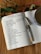 An open notebook displays a handwritten recipe for dark chocolate and cherry Christmas pudding. A pen rests on the notebook, which is placed on a wooden surface. Nearby, a cup filled with a brown beverage and some green foliage add to the composition.