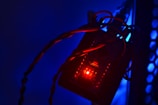 A close-up view of an electronic circuit board illuminated with a red LED light. Twisted wires are connected to the circuit board, and the background is bathed in blue lighting. There is a honeycomb-like pattern visible on the right side, possibly part of a protective casing.